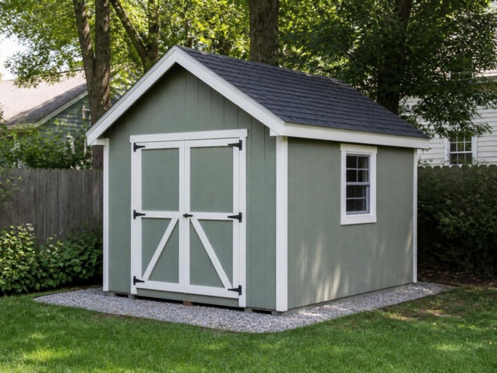Sage green custom storage shed with white trim and braced double doors on a gravel pad in a backyard